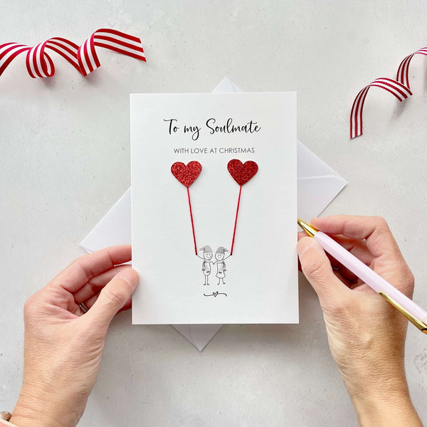 A Christmas card featuring a minimalist design with two figures wearing santa hats holding red glitter heart balloons. The text at the top reads: ‘To my Soulmate, with love at Christmas’. The card is placed on a white background with a white envelope partially visible behind it. A hand is seen holding the card, while another hand holds a pink pen. A red and white striped ribbon is positioned in the top corners of the image for decoration.