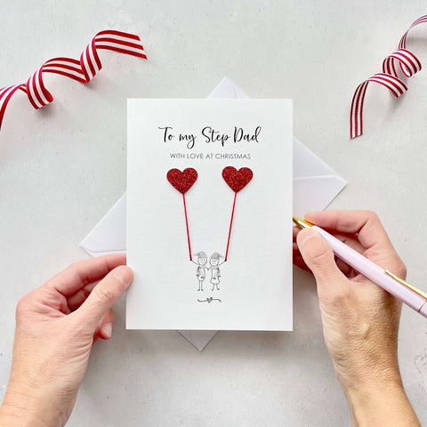 A Christmas card featuring a minimalist design with two figures wearing santa hats holding red glitter heart balloons. The text at the top reads: ‘To my Step-Dad, with love at Christmas’. The card is placed on a white background with a white envelope partially visible behind it. A hand is seen holding the card, while another hand holds a pink pen. A red and white striped ribbon is positioned in the top corners of the image for decoration.