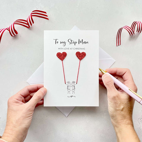 A Christmas card featuring a minimalist design with two figures wearing santa hats holding red glitter heart balloons. The text at the top reads: ‘To my Step-Mum, with love at Christmas’. The card is placed on a white background with a white envelope partially visible behind it. A hand is seen holding the card, while another hand holds a pink pen. A red and white striped ribbon is positioned in the top corners of the image for decoration.