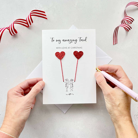 A Christmas card featuring a minimalist design with two figures wearing santa hats holding red glitter heart balloons. The text at the top reads: ‘To my amazing Taid, With Love at Christmas’. The card is placed on a white background with a white envelope partially visible behind it. A hand is seen holding the card, while another hand holds a pink pen. A red and white striped ribbon is positioned in the top corners of the image for decoration.