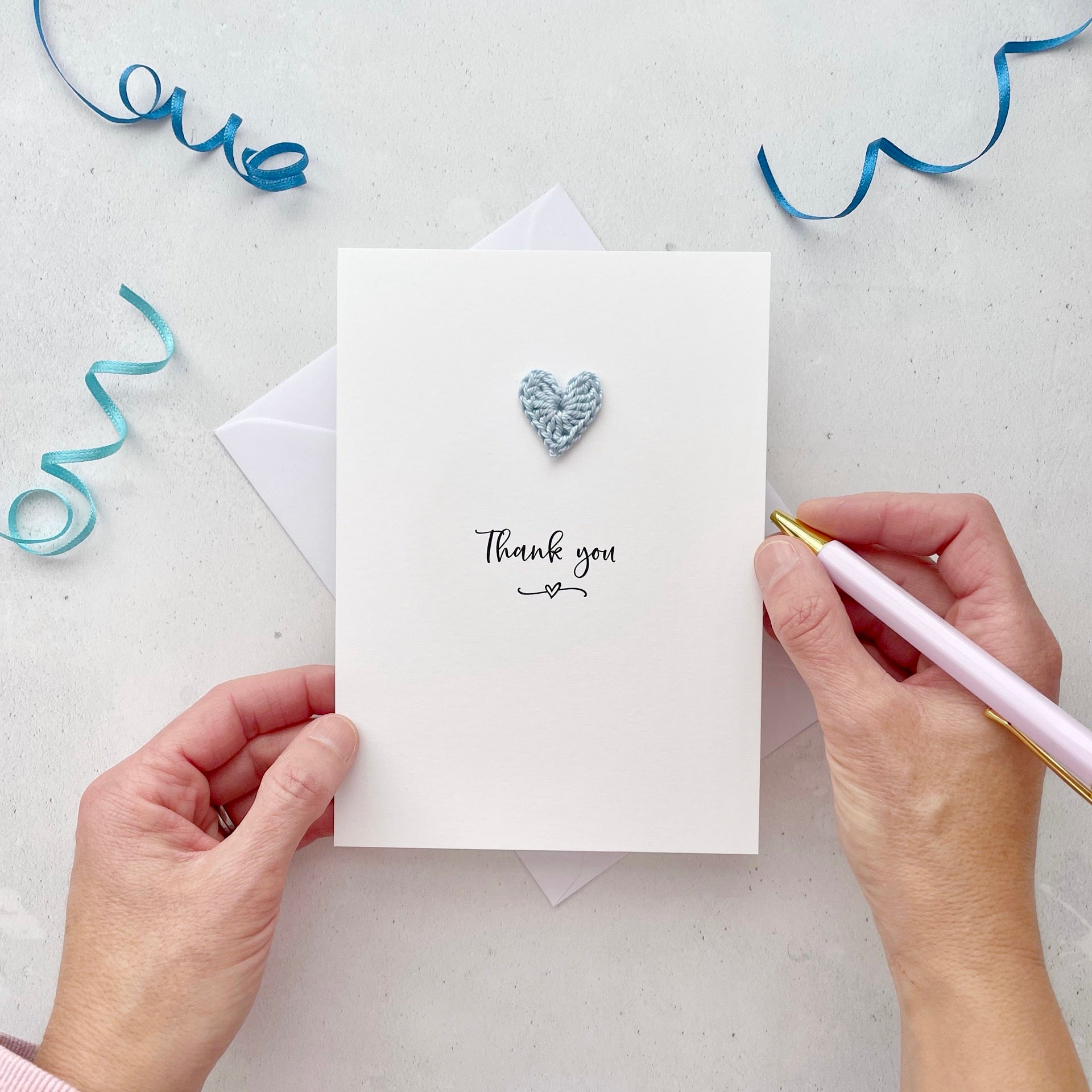 A white portrait-style card featuring the words ‘Thank you’ in elegant black cursive at the center. Below the text, a small black squiggle and heart design adds a playful touch. Above the words, a handmade pale blue crochet heart is carefully attached, providing a pop of colour and texture.