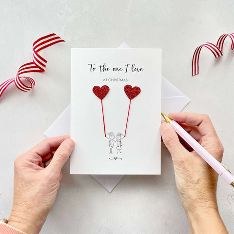 A Christmas card featuring a minimalist design with two figures wearing santa hats holding red glitter heart balloons. The text at the top reads: ‘To the one I love, at Christmas’. The card is placed on a white background with a white envelope partially visible behind it. A hand is seen holding the card, while another hand holds a pink pen. A red and white striped ribbon is positioned in the top corners of the image for decoration.