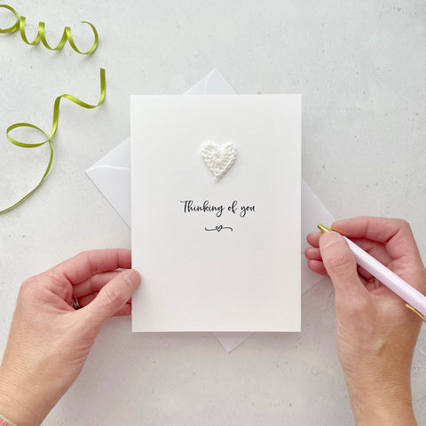 A white portrait-style card featuring the words ‘Thinking of you’ in elegant black cursive at the center. Below the text, a small black squiggle and heart design adds a playful touch. Above the words, a handmade ivory crochet heart is carefully attached, providing a pop of colour and texture.