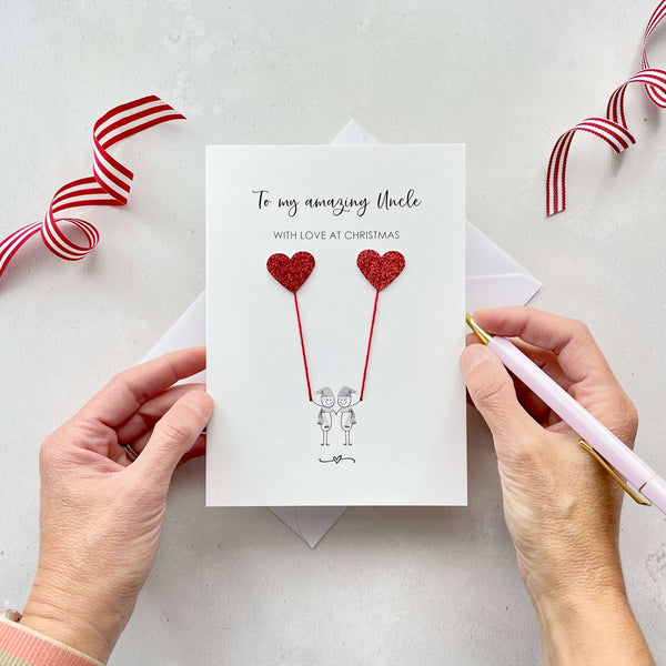 A Christmas card featuring a minimalist design with two figures wearing santa hats holding red glitter heart balloons. The text at the top reads: "To my amazing Uncle, With Love at Christmas." The card is placed on a white background with a white envelope partially visible behind it. A hand is seen holding the card, while another hand holds a pink pen. A red and white striped ribbon is positioned in the top corners of the image for decoration.