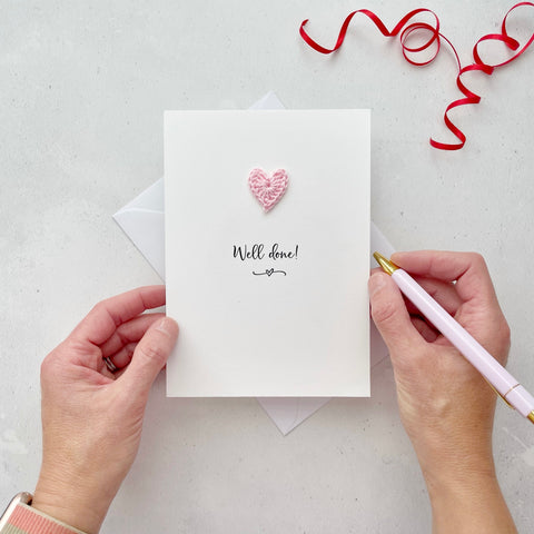 A white portrait-style card featuring the words ’Well done!’ in elegant black cursive at the center. Below the text, a small black squiggle and heart design adds a playful touch. Above the words, a handmade pale pink crochet heart is carefully attached, providing a pop of colour and texture.