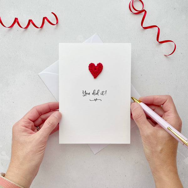 A white portrait-style card featuring the words ’You did it!’ in elegant black cursive at the center. Below the text, a small black squiggle and heart design adds a playful touch. Above the words, a handmade red crochet heart is carefully attached, providing a pop of colour and texture.