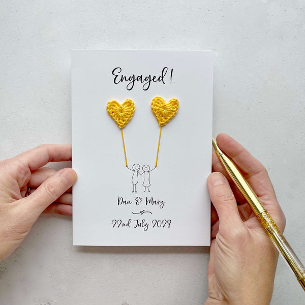 The image shows a beautiful handmade engagement card with little yellow crochet hearts, these are attached by cotton yarn, resembling balloons being held by a hand-drawn couple underneath. The text on the card reads “Engaged! at the top, followed by the couples names and engagement date at the bottom. The card is set on a light background, and the person's hands are seen holding a pen and gently holding the card, giving the image a sense of care and personalisation.