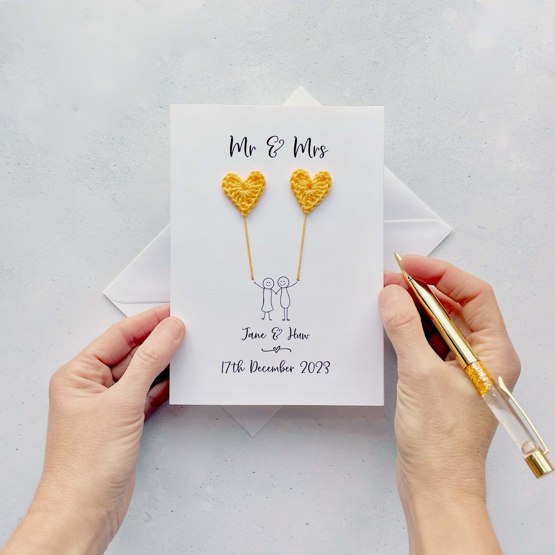 The image shows a beautiful personalised wedding card with little yellow crochet hearts, these are attached by cotton yarn, resembling balloons being held by a hand-drawn couple underneath. The text on the card reads 'Mr & Mrs' at the top, followed by the couples names and wedding date at the bottom.