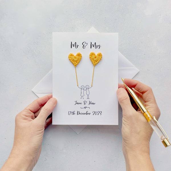 The image shows a beautiful personalised wedding card with little yellow crochet hearts, these are attached by cotton yarn, resembling balloons being held by a hand-drawn couple underneath. The text on the card reads 'Mr & Mrs' at the top, followed by the couples names and wedding date at the bottom.