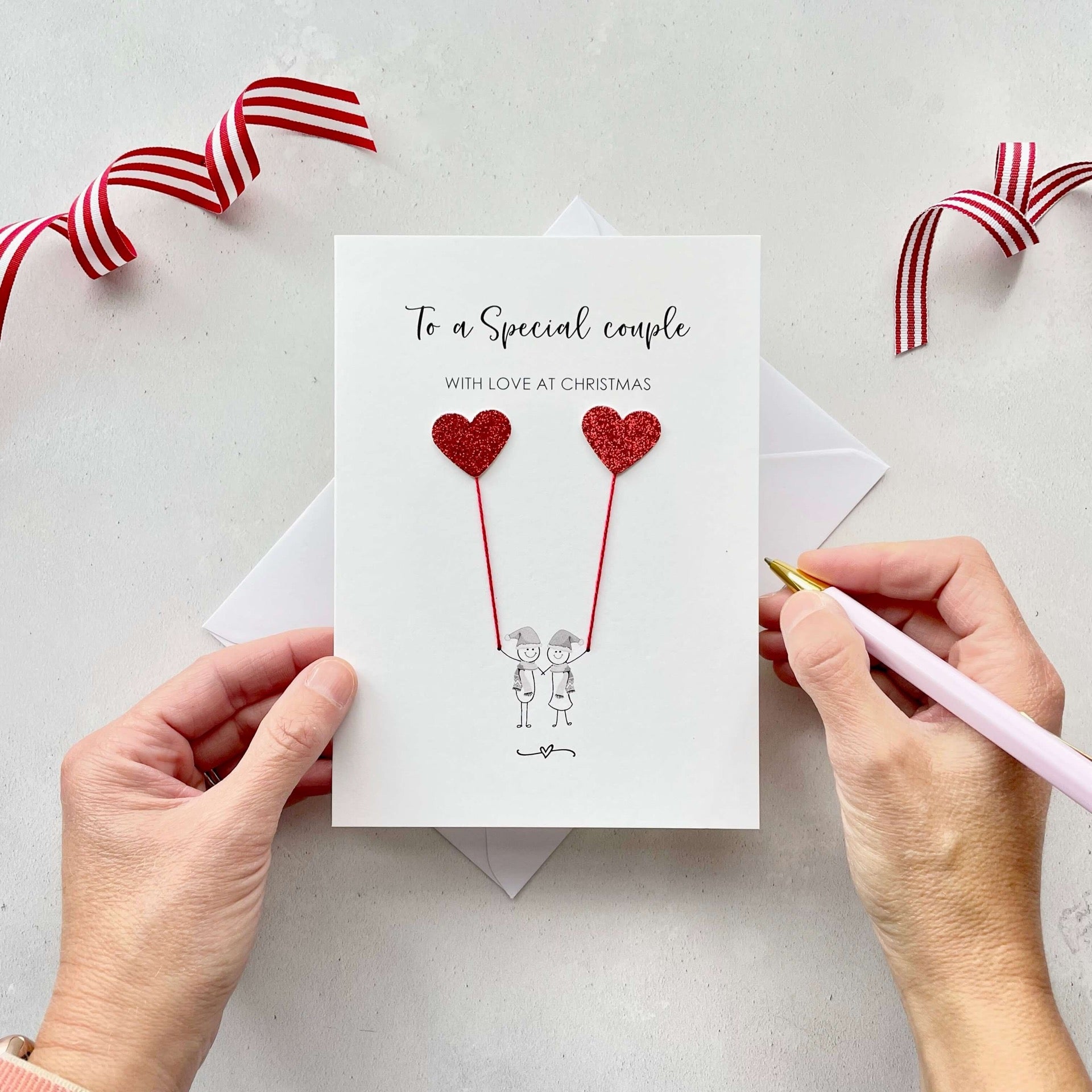 A Christmas card featuring a minimalist design with two figures wearing santa hats holding red glitter heart balloons. The text at the top reads: "To a Special Couple, With Love at Christmas." The card is placed on a white background with a white envelope partially visible behind it. A hand is seen holding the card, while another hand holds a pink pen. A red and white striped ribbon is positioned in the top corners of the image for decoration.