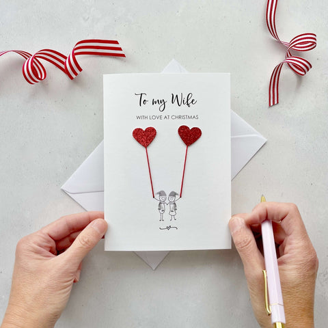 A Christmas card featuring a minimalist design with two figures wearing santa hats holding red glitter heart balloons. The text at the top reads: "To my Wife, With Love at Christmas." The card is placed on a white background with a white envelope partially visible behind it. A hand is seen holding the card, while another hand holds a pink pen. A red and white striped ribbon is positioned in the top corners of the image for decoration.