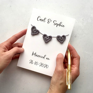 A white personalised wedding card with  'Carl & Sophie" printed in black cursive text at the top of the card. Below the names are 3 mini grey crochet hearts strung onto the card like bunting. It reads 'Married on' and 24-10-2020' at the bottom of the card. 